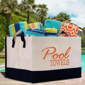 May include: A white canvas tote bag with navy blue trim and handles, filled with rolled pool towels. The bag has the words "Pool TOWELS" in orange script. The towels are various colours and patterns, including stripes and geometric designs.