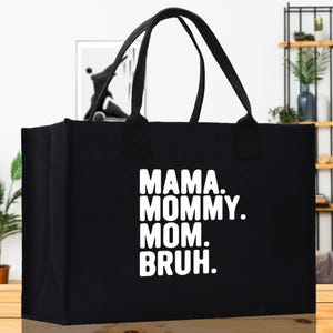 May include: Black tote bag with white text that reads "MAMA. MOMMY. MOM. BRUH." The bag has two black handles and is rectangular in shape. The background includes a shelf with plants and a framed picture.