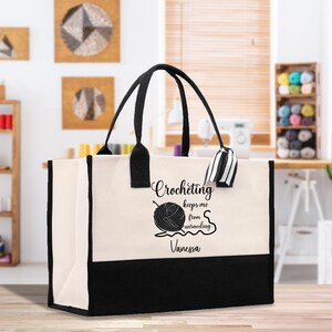 May include: A large tote bag with black handles and a black bottom panel. The bag is cream-colored with the words "Crocheting keeps me from unraveling" and a yarn ball graphic. The name "Vanessa" is below the text. A black and white striped tag hangs from the handle.