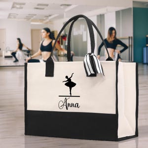 Può includere: Una borsa tote in tela con finiture e manici neri. La borsa presenta una silhouette nera di una ballerina e il nome "Anna" in corsivo. Un nastro a righe bianche e nere è attaccato al manico.