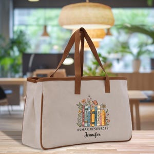 May include: A beige canvas tote bag with brown leather handles and trim. The bag features a colorful floral design with a stack of books and the words "HUMAN RESOURCES" and a name. The bag is designed for carrying books and other items.
