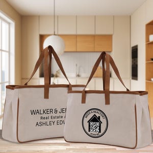 Op de afbeelding: Twee canvas draagtassen met bruine randen en handvatten. Eén tas heeft de tekst "WALKER & JENNIFER Real Estate / ASHLEY EDWARDS". De andere tas heeft een cirkelvormig logo met een huis en de tekst "NEED A REALTOR? SCAN ME".