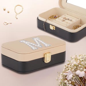 May include: A two-tone jewelry box with a beige top and black base, featuring a large, sparkling letter "M" on the lid. The box is open, revealing compartments and jewelry. A gold clasp secures the box. Pearls and other jewelry are scattered around.