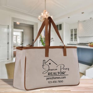 Könnte beinhalten: Eine beigefarbene Canvas-Tragetasche mit braunen Ledergriffen und -besatz. Die Tasche trägt den Namen "Jessica Perez REALTOR" und Kontaktinformationen in schwarzer Schrift. Die Tasche ist zum Tragen von Gegenständen bestimmt.