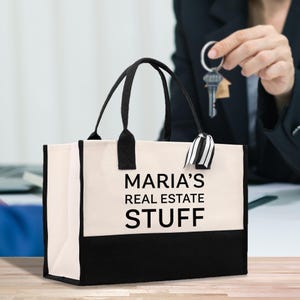 Può includere: Una borsa tote in tela color crema con manici neri e base nera. La borsa presenta la scritta "MARIA'S REAL ESTATE STUFF" in nero. Una persona tiene delle chiavi sullo sfondo.