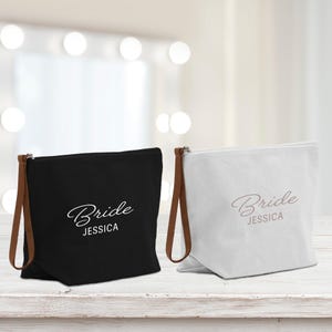 Op de afbeelding: Twee canvas cosmetische tassen, één zwart en één wit, met bruine leren handvatten. Elke tas is bedrukt met het woord "Bride" en een naam in een scriptlettertype. De tassen staan op een wit houten oppervlak.
