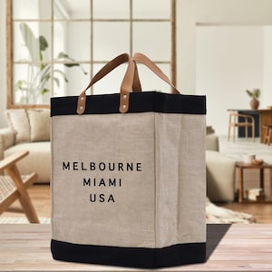 May include: A rectangular tote bag with a beige body, black top and bottom trim, and tan leather handles. The bag features the text "MELBOURNE MIAMI USA" in black. The bag is sitting on a wooden surface.