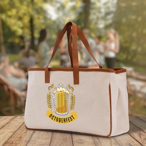 May include: Cream-coloured tote bag with brown leather handles and trim. The bag features a colourful graphic of a beer stein with the word "OCTOBERFEST" on a yellow banner. The bag is sitting on a wooden surface.