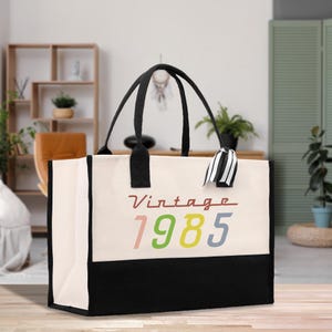 May include: A canvas tote bag with black handles and a black bottom panel. The bag features the text "Vintage 1985" in a colorful font. A black and white striped ribbon is attached to one of the handles.