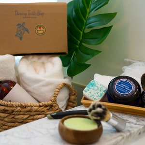 May include: A brown cardboard box with the text "Dancing Turtle Shop" printed on it. The box is filled with white towels, a brown bottle with a gold pump, and a white bathrobe. The box is sitting on a white countertop next to a tray of shaving supplies, including a shaving cream jar, a shaving brush, and a bar of soap.
