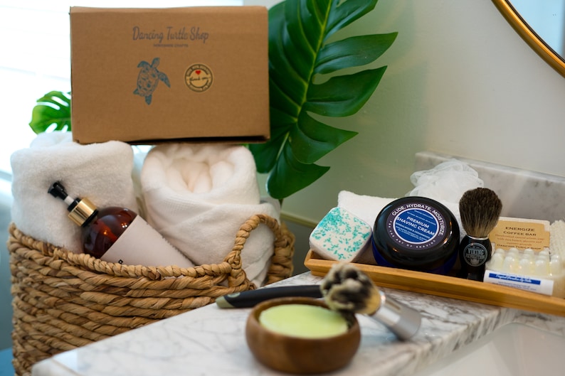 May include: A brown cardboard box with the text "Dancing Turtle Shop" printed on it. The box is filled with white towels, a brown bottle with a gold pump, and a white bathrobe. The box is sitting on a white countertop next to a tray of shaving supplies, including a shaving cream jar, a shaving brush, and a bar of soap.