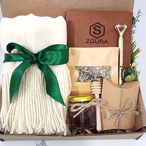 May include: A gift box filled with a white scarf with green ribbon, a brown leather journal with the text "ZGURA" and a gold pen, a small jar of honey, a wooden honey dipper, a small bag of tea, and a brown paper bag with a string tie.