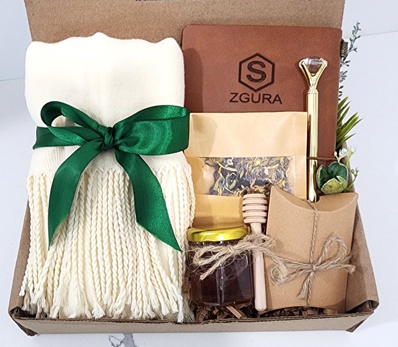 May include: A gift box filled with a white scarf with green ribbon, a brown leather journal with the text "ZGURA" and a gold pen, a small jar of honey, a wooden honey dipper, a small bag of tea, and a brown paper bag with a string tie.