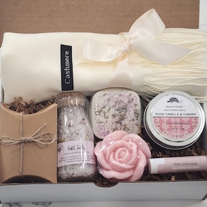 May include: A gift box filled with self-care items, including a white cashmere scarf, a brown paper bag, a clear glass jar of bath salts labelled "Bath Salts Vanilla Bean Soak", a white soap bar, a pink rose-shaped soap, a small tin candle labelled "Rose Vanilla & Cherry", and a pink lip balm labelled "Pink Lemonade".