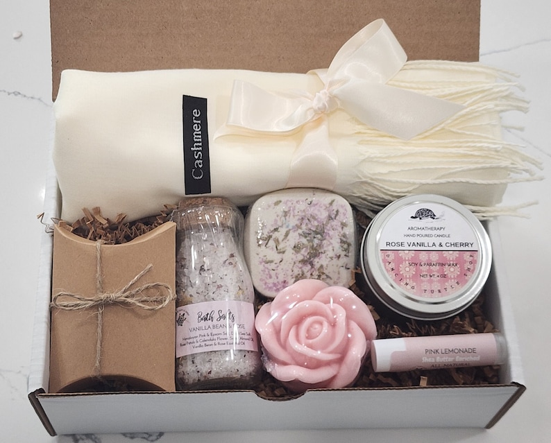 May include: A gift box filled with self-care items, including a white cashmere scarf, a brown paper bag, a clear glass jar of bath salts labelled "Bath Salts Vanilla Bean Soak", a white soap bar, a pink rose-shaped soap, a small tin candle labelled "Rose Vanilla & Cherry", and a pink lip balm labelled "Pink Lemonade".