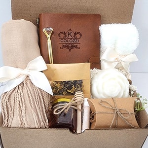 May include: A gift box filled with a variety of items, including a brown leather journal with the text "K COLOZZI" engraved on the cover, a white towel, a white soap, a jar of honey, a small bag of tea, a pen with a diamond-shaped top, and a beige scarf with a fringe.
