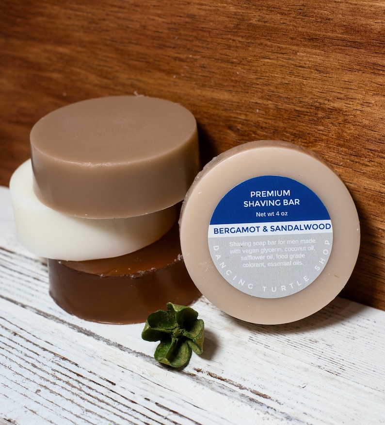 May include: A stack of four round, brown, white and beige shaving soap bars. The top bar has a blue label that reads "Premium Shaving Bar, Net wt 4 oz, Bergamot & Sandalwood, Dancing Turtle Shop".