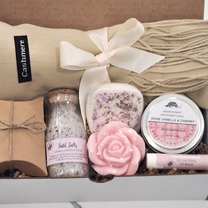 May include: A gift box filled with self-care items, including a cashmere scarf, bath salts, a rose-shaped soap, a candle, and lip balm. The box is decorated with brown paper and a white ribbon.