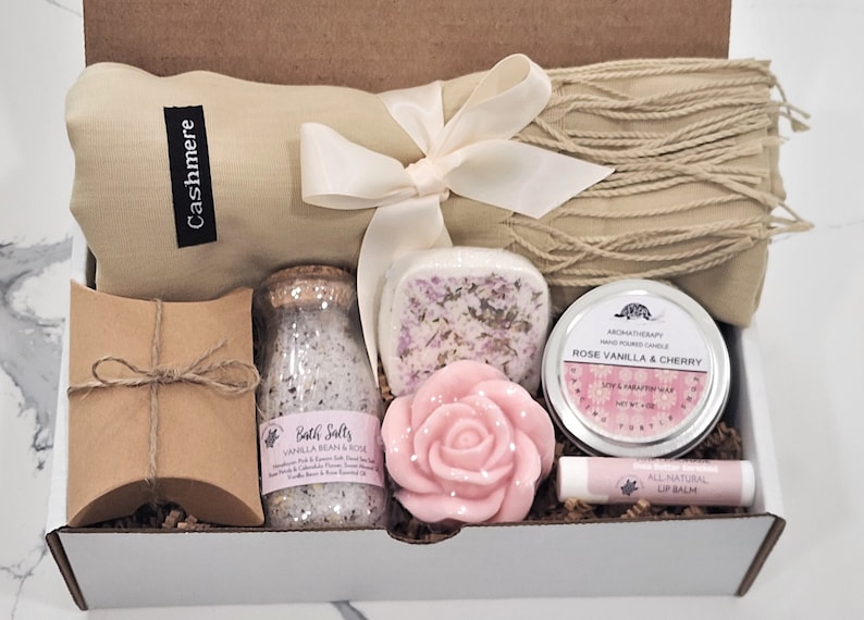 May include: A gift box filled with self-care items, including a cashmere scarf, bath salts, a rose-shaped soap, a candle, and lip balm. The box is decorated with brown paper and a white ribbon.