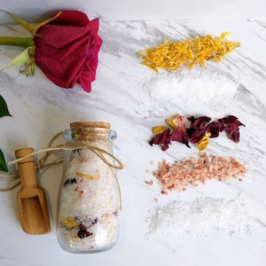 May include: A glass jar filled with bath salts, rose petals, and dried citrus. The jar is tied with twine and has a cork lid. A wooden scoop is next to the jar. The bath salts are surrounded by dried rose petals, dried citrus, and white bath salts.