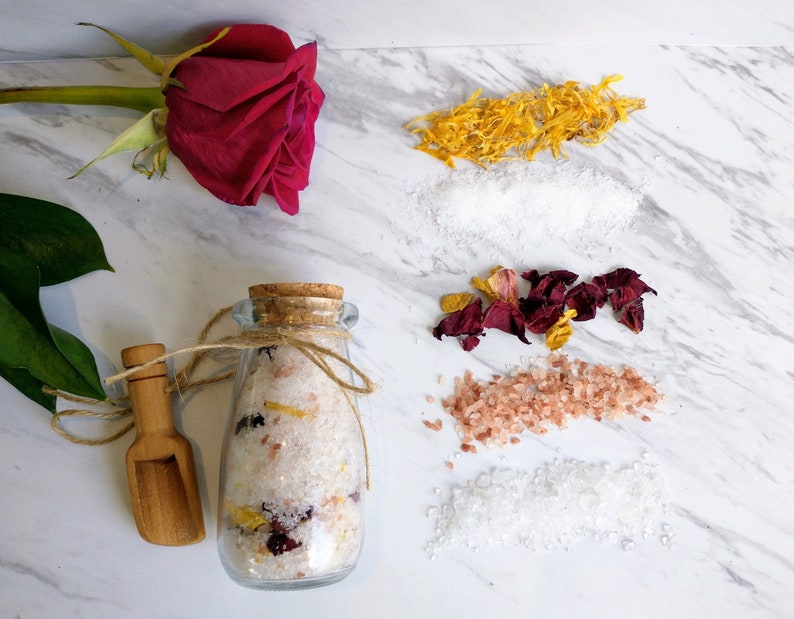 May include: A glass jar filled with bath salts, rose petals, and dried citrus. The jar is tied with twine and has a cork lid. A wooden scoop is next to the jar. The bath salts are surrounded by dried rose petals, dried citrus, and white bath salts.