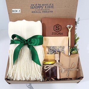 May include: A gift box labeled "BOX 2" with the text "one of the secrets of a HAPPY LIFE is continuous SMALL TREATS". The box contains a white scarf with green fringe and a green ribbon, a brown leather journal with the text "ZGURA" and a gold pen, a small jar of honey, a wooden honey dipper, a small bag of loose leaf tea, and a brown paper envelope.