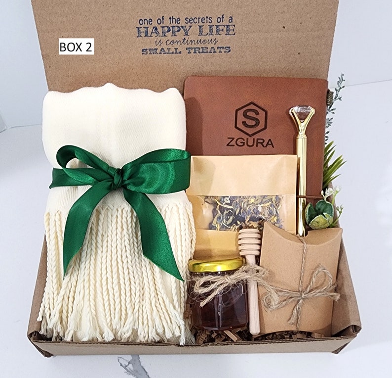 May include: A gift box labeled "BOX 2" with the text "one of the secrets of a HAPPY LIFE is continuous SMALL TREATS". The box contains a white scarf with green fringe and a green ribbon, a brown leather journal with the text "ZGURA" and a gold pen, a small jar of honey, a wooden honey dipper, a small bag of loose leaf tea, and a brown paper envelope.