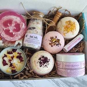 K&ouml;nnte beinhalten: Eine Geschenkbox gef&uuml;llt mit Bade- und K&ouml;rperpflegeprodukten, darunter Badesalz, Badebomben, eine Kerze, ein Lippenbalsam und K&ouml;rperbutter. Die Produkte sind alle in Rosat&ouml;nen, Wei&szlig; und Gelb gehalten, und die Box ist mit braunem Papier ausgekleidet.