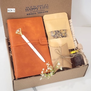 May include: A brown gift box with the text "one of the secrets of a HAPPY LIFE is continuous SMALL TREATS" printed on the top. The box contains a brown leather journal, a white pen, a small bag of dried flowers, a small brown paper bag, a small jar of honey, and a wooden honey dipper.