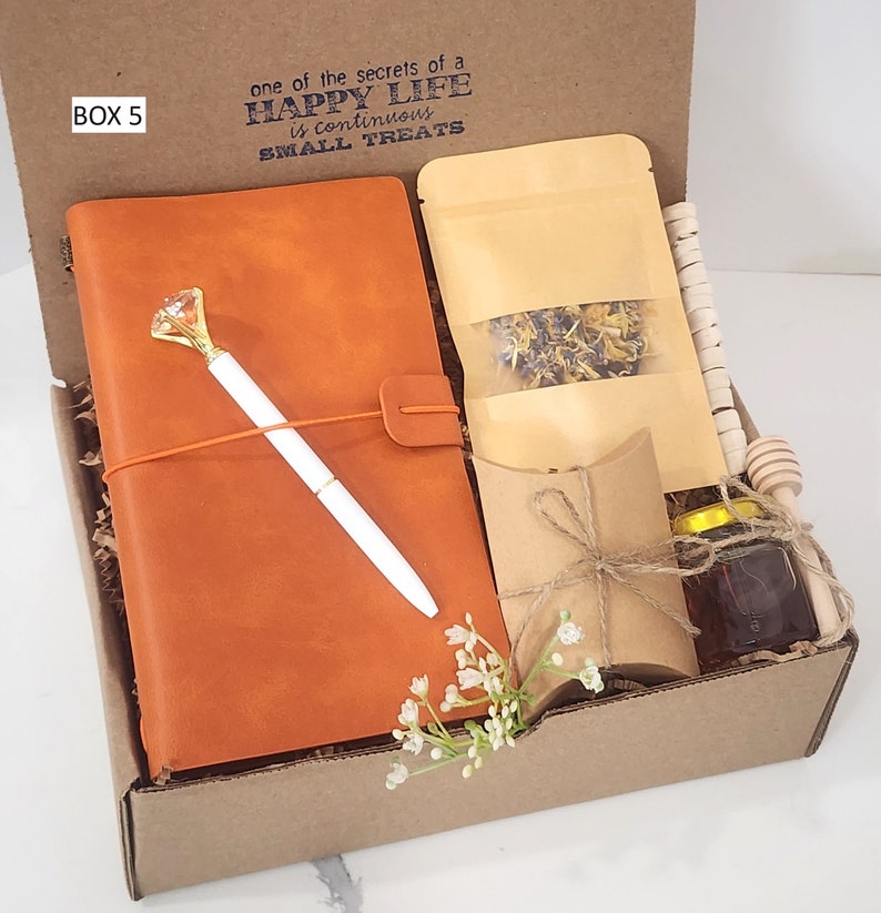 May include: A brown gift box with the text "one of the secrets of a HAPPY LIFE is continuous SMALL TREATS" printed on the top. The box contains a brown leather journal, a white pen, a small bag of dried flowers, a small brown paper bag, a small jar of honey, and a wooden honey dipper.