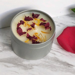 May include: A silver tin candle with a white wax top. The wax is decorated with dried red and yellow flower petals.