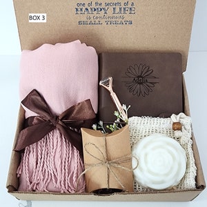 May include: A gift box labeled "BOX 3" with a pink scarf, a brown journal with the name "Adelia" engraved on it, a small brown pillow, and a white soap in a mesh bag. The box lid has the text "one of the secrets of a HAPPY LIFE is continuous SMALL TREATS".