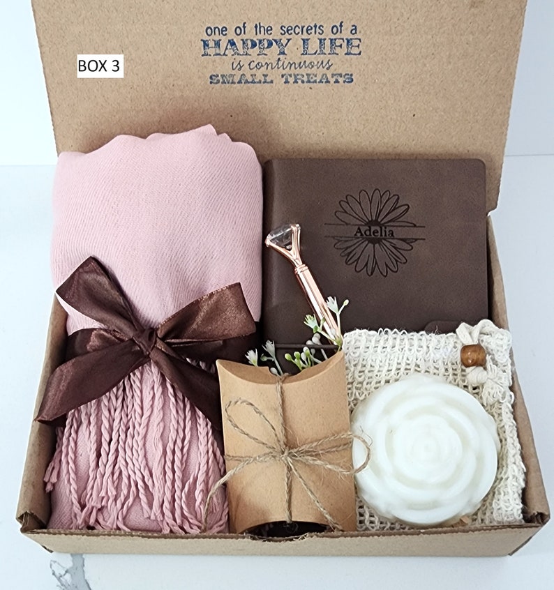 May include: A gift box labeled "BOX 3" with a pink scarf, a brown journal with the name "Adelia" engraved on it, a small brown pillow, and a white soap in a mesh bag. The box lid has the text "one of the secrets of a HAPPY LIFE is continuous SMALL TREATS".
