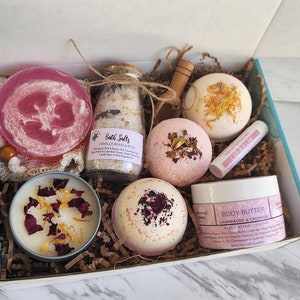 K&ouml;nnte beinhalten: Eine Geschenkbox gef&uuml;llt mit Bade- und K&ouml;rperpflegeprodukten. Die Box enth&auml;lt Badebomben, ein Glas Badesalz, eine Kerze, ein Luffa, einen Lippenbalsam und ein Glas K&ouml;rperbutter. Die Produkte sind alle in Rosa- und Wei&szlig;t&ouml;nen gehalten.