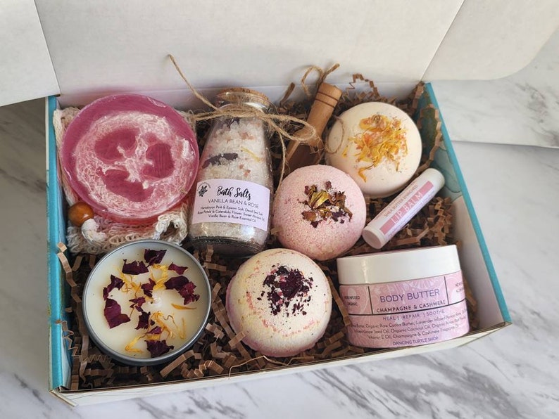 K&ouml;nnte beinhalten: Eine Geschenkbox gef&uuml;llt mit Bade- und K&ouml;rperpflegeprodukten. Die Box enth&auml;lt Badebomben, ein Glas Badesalz, eine Kerze, ein Luffa, einen Lippenbalsam und ein Glas K&ouml;rperbutter. Die Produkte sind alle in Rosa- und Wei&szlig;t&ouml;nen gehalten.