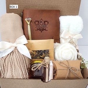 May include: A brown gift box labeled "BOX 1" filled with various items, including a brown leather journal with a personalized inscription, a white towel, a brown scarf, a jar of honey, a small bag of tea, a white soap, a wooden honey dipper, and a brown paper bag.