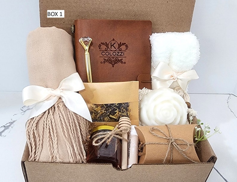 May include: A brown gift box labeled "BOX 1" filled with various items, including a brown leather journal with a personalized inscription, a white towel, a brown scarf, a jar of honey, a small bag of tea, a white soap, a wooden honey dipper, and a brown paper bag.