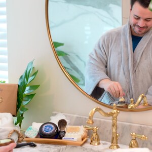May include: A man in a grey dressing gown stands in front of a bathroom mirror, using a shaving brush. The mirror is round with a gold frame. The bathroom counter is white with gold taps. There are various grooming products on the counter, including a shaving brush, shaving cream, and soap.