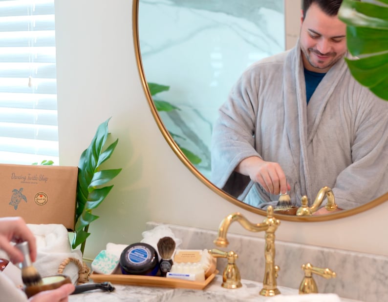 May include: A man in a grey dressing gown stands in front of a bathroom mirror, using a shaving brush. The mirror is round with a gold frame. The bathroom counter is white with gold taps. There are various grooming products on the counter, including a shaving brush, shaving cream, and soap.