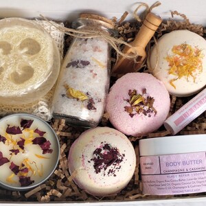K&ouml;nnte beinhalten: Eine Geschenkbox gef&uuml;llt mit Bade- und K&ouml;rperpflegeprodukten, darunter Badebomben, eine Kerze, K&ouml;rperbutter, Badesalz, ein Luffa-Schwamm und Lippenbalsam. Die Produkte sind alle mit getrockneten Blumen und Bl&uuml;tenbl&auml;ttern verziert.