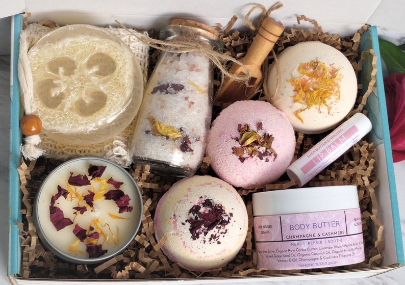 K&ouml;nnte beinhalten: Eine Geschenkbox gef&uuml;llt mit Bade- und K&ouml;rperpflegeprodukten, darunter Badebomben, eine Kerze, K&ouml;rperbutter, Badesalz, ein Luffa-Schwamm und Lippenbalsam. Die Produkte sind alle mit getrockneten Blumen und Bl&uuml;tenbl&auml;ttern verziert.