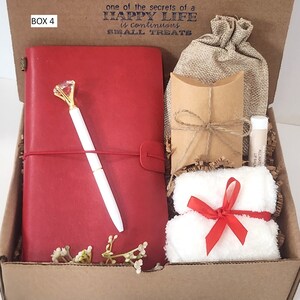 May include: A gift box filled with items including a red journal with a pen, a small brown bag, a white towel with a red ribbon, and a small tube of lip balm. The box is labeled "BOX 4" and has the text "one of the secrets of a HAPPY LIFE is continuous SMALL TREATS" printed on the top.