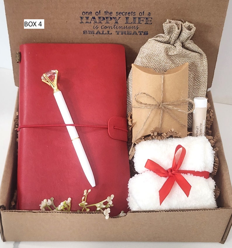 May include: A gift box filled with items including a red journal with a pen, a small brown bag, a white towel with a red ribbon, and a small tube of lip balm. The box is labeled "BOX 4" and has the text "one of the secrets of a HAPPY LIFE is continuous SMALL TREATS" printed on the top.