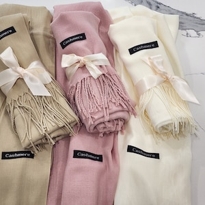 May include: Three cashmere scarves in beige, pink, and white, each tied with a satin ribbon and featuring a black tag that reads "Cashmere".