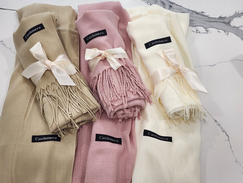 May include: Three cashmere scarves in beige, pink, and white, each tied with a satin ribbon and featuring a black tag that reads "Cashmere".
