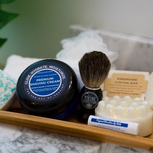 May include: A bamboo tray containing a selection of men's grooming products, including a shaving cream jar, a shaving brush, a coffee bar, a soap bar, and a lip balm. The shaving cream jar is labelled "Glide Hydrate Moisturize Premium Shaving Cream Sandalwood & Cedarwood". The shaving brush is labelled "Viking Revolution". The coffee bar is labelled "Energize Coffee Bar". The soap bar is white and has a textured surface. The lip balm is labelled "Tropical Paradise Lip Balm".