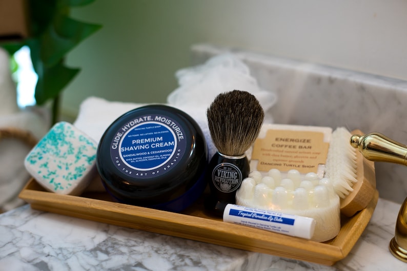 May include: A bamboo tray containing a selection of men's grooming products, including a shaving cream jar, a shaving brush, a coffee bar, a soap bar, and a lip balm. The shaving cream jar is labelled "Glide Hydrate Moisturize Premium Shaving Cream Sandalwood & Cedarwood". The shaving brush is labelled "Viking Revolution". The coffee bar is labelled "Energize Coffee Bar". The soap bar is white and has a textured surface. The lip balm is labelled "Tropical Paradise Lip Balm".