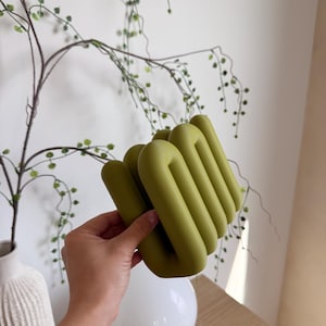May include: A matte olive green decorative object featuring a series of interconnected, rounded, tubular loops arranged in a vertical, accordion-like formation. The structure has a smooth, uniform surface texture and a solid, continuous design.