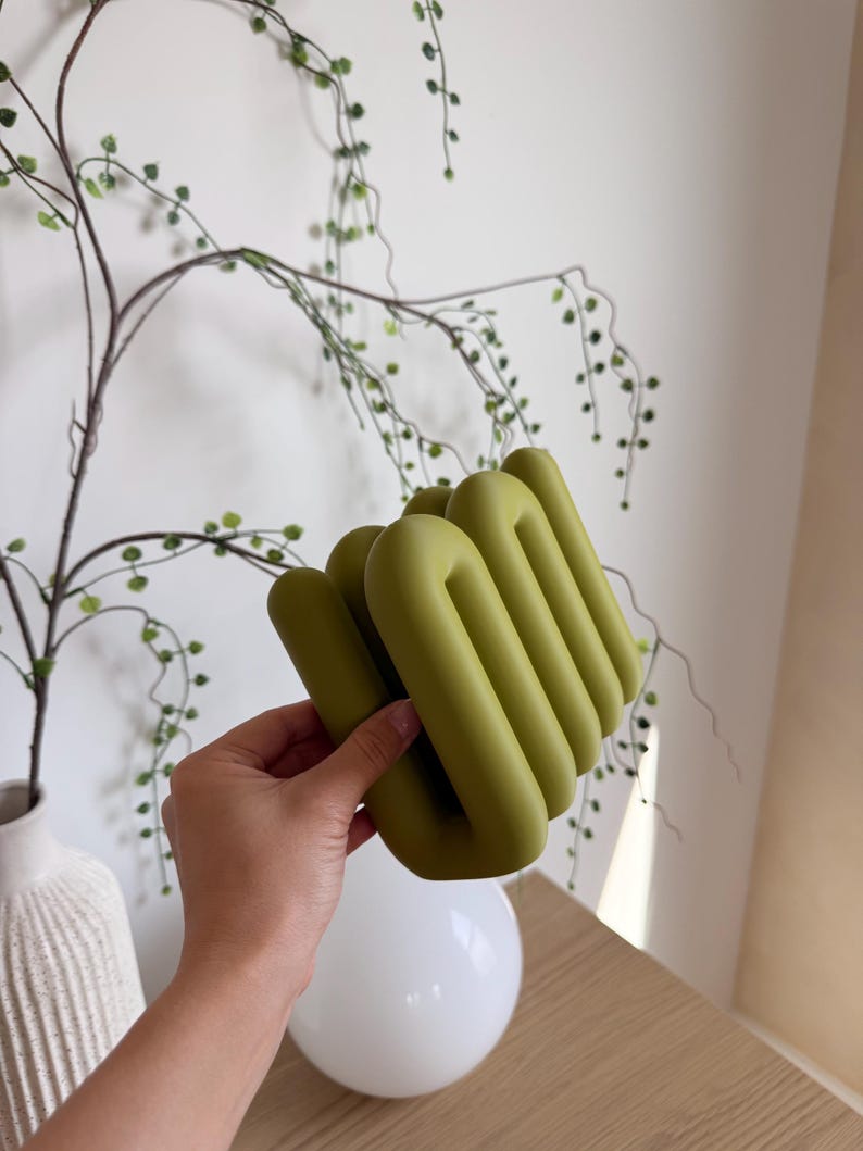 May include: A matte olive green decorative object featuring a series of interconnected, rounded, tubular loops arranged in a vertical, accordion-like formation. The structure has a smooth, uniform surface texture and a solid, continuous design.
