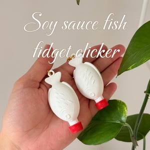 May include: Two white, fish-shaped fidget clickers with red caps are held in a hand. The text "Soy sauce fish fidget clicker" is above the fidget toys. The background includes green leaves and a white wall.